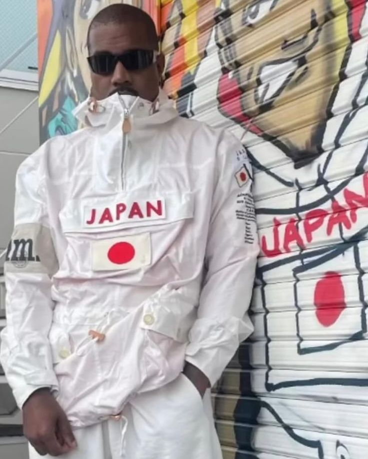 a man standing in front of a wall with graffiti on it and wearing white clothing