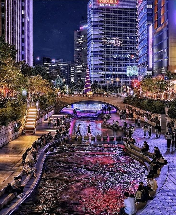 people are sitting on the edge of a river in front of tall buildings at night