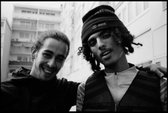 two young men standing next to each other in front of tall buildings and smiling at the camera