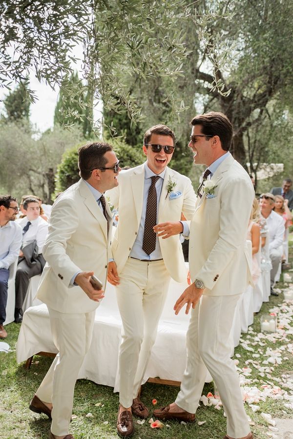 three men in white suits standing next to each other at a wedding ceremony under an olive tree