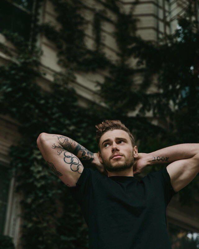 a man with tattoos standing in front of a building and holding his arms behind his head