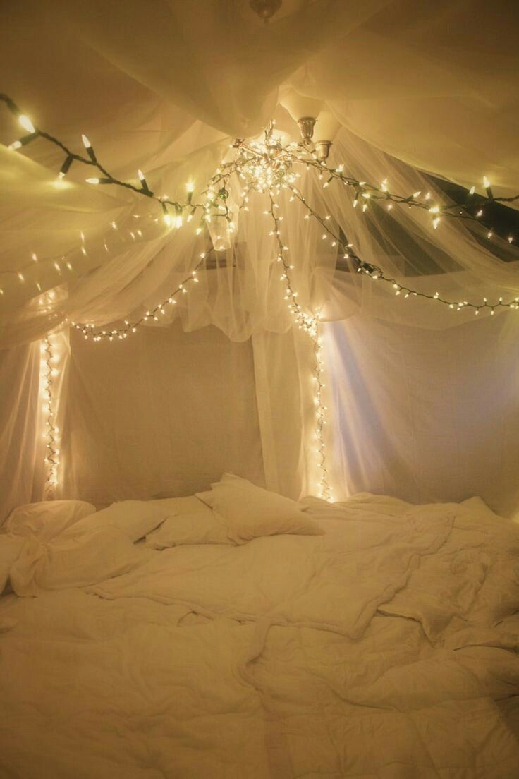 a bed covered in white sheets with lights on the ceiling and curtains hanging over it