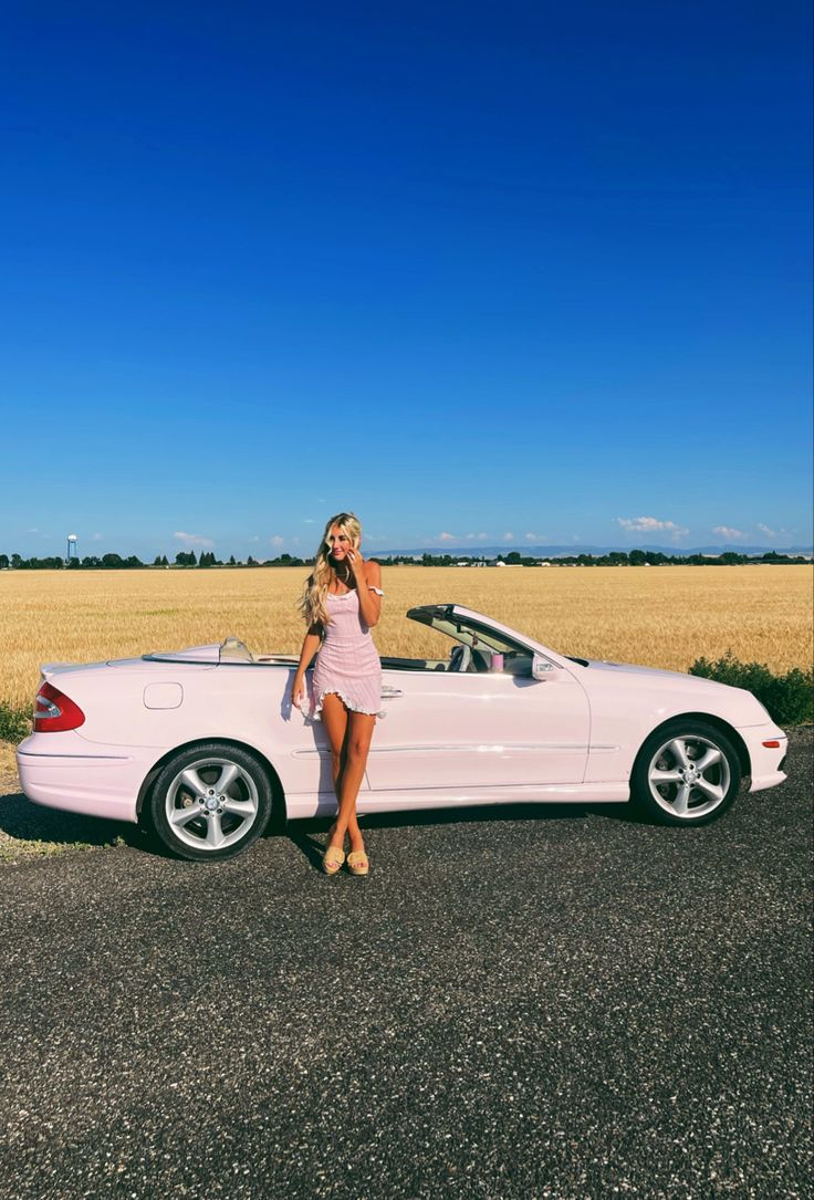 a woman standing next to a pink car in the middle of a road with wheat fields behind her