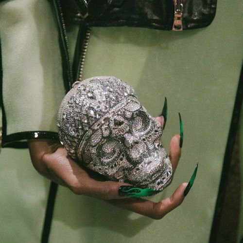 a hand holding a skull shaped purse with green nails on it's fingers and black leather bag in the background