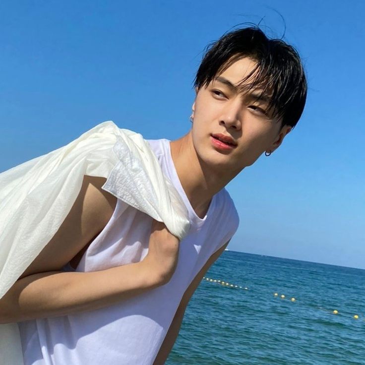 a young man standing on top of a beach next to the ocean with his shirt over his shoulder