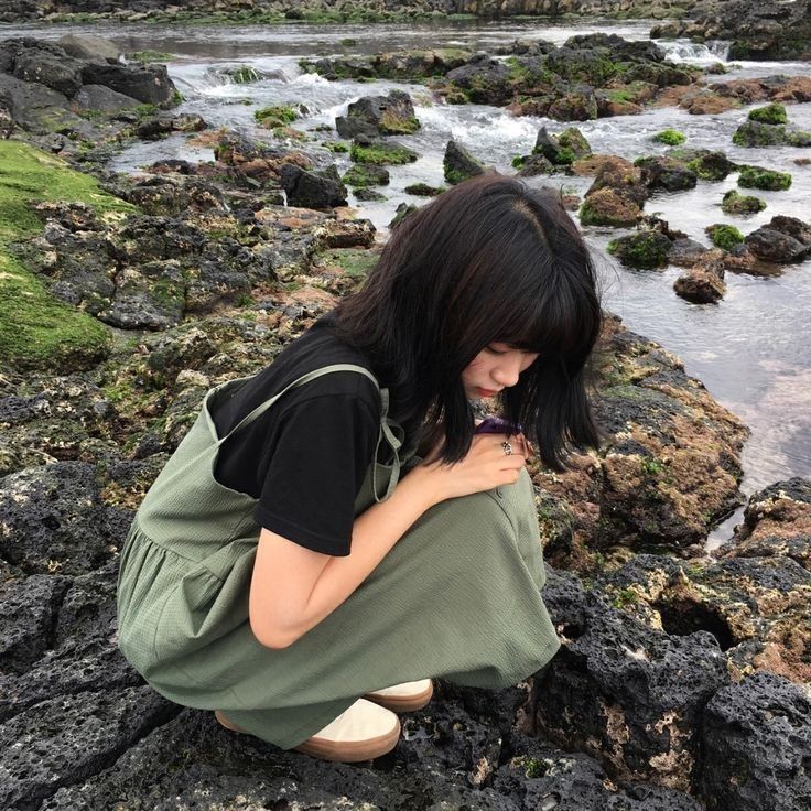 a woman crouches on rocks and looks at her cell phone in front of the water