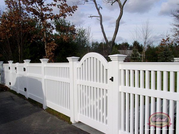 a white picket fence in front of a driveway with trees and bushes behind it on a cloudy day