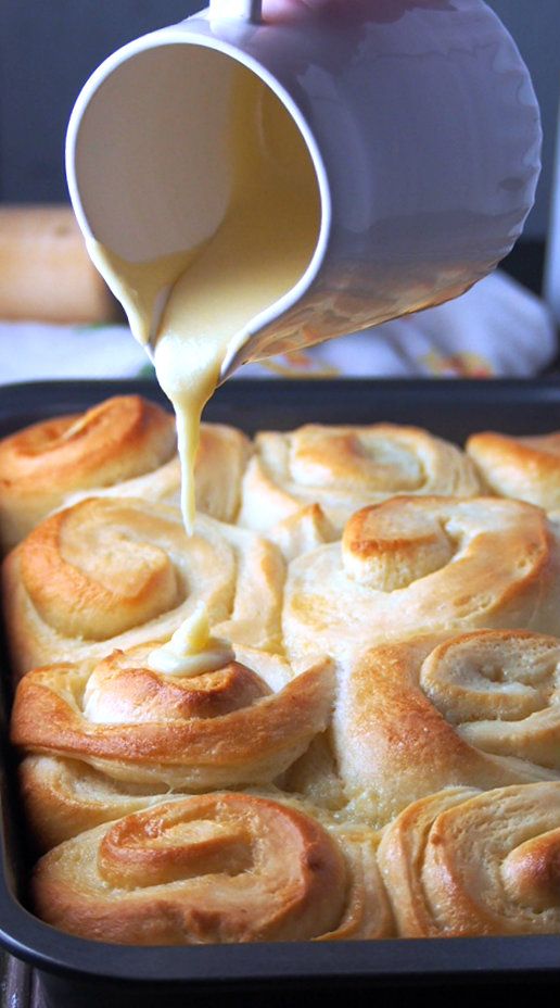 a person pouring sauce on top of a baked pastry in a baking pan with cinnamon rolls