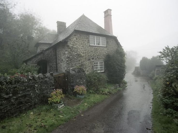 an old stone house in the middle of a foggy road with flowers and bushes on either side