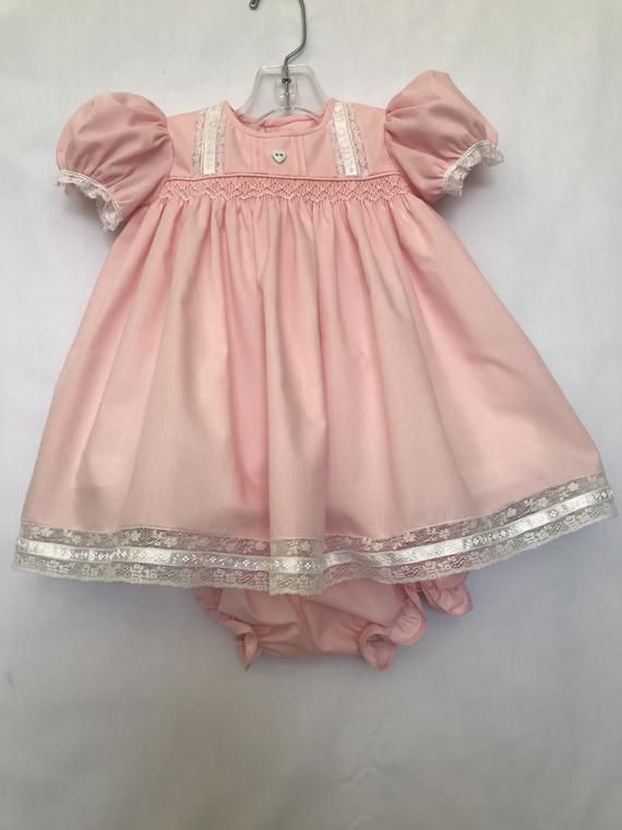 This size 3 mo. dress is made of pink, poly/cotton fabric. Lace insertions and edgings are on the bodice, sleeves and dress bottom. The dress is lined with white polyester cooton. A white heart button is added to the bodice and the back is also fastened with 2 heart buttons. The body of the dress Cotton Baptism Dress With Lining, Cotton Short Sleeve Dress For Baptism, Fitted Pink Cotton Dress, Cute Pink Dresses With Buttons, Cute Pink Dress With Buttons, Fitted Pink Dress With Buttons, Cute Pink Lined Dress, Pink Lace Trim Dress For Baptism, Pink Cotton Smock Dress