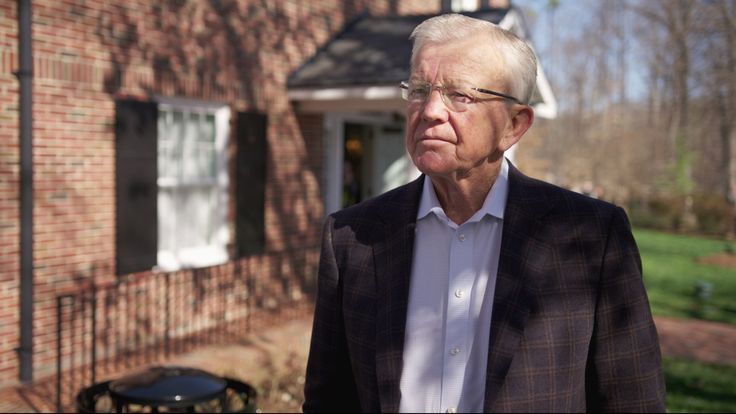 an older man standing in front of a red brick house wearing glasses and a blazer