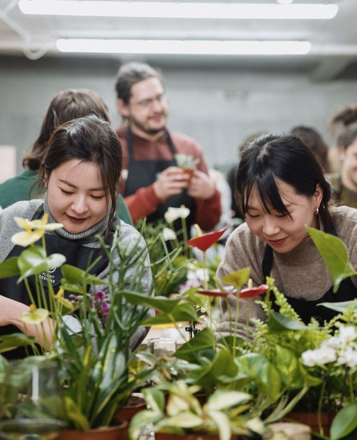 two women looking at plants in pots on a table with other people behind them and smiling