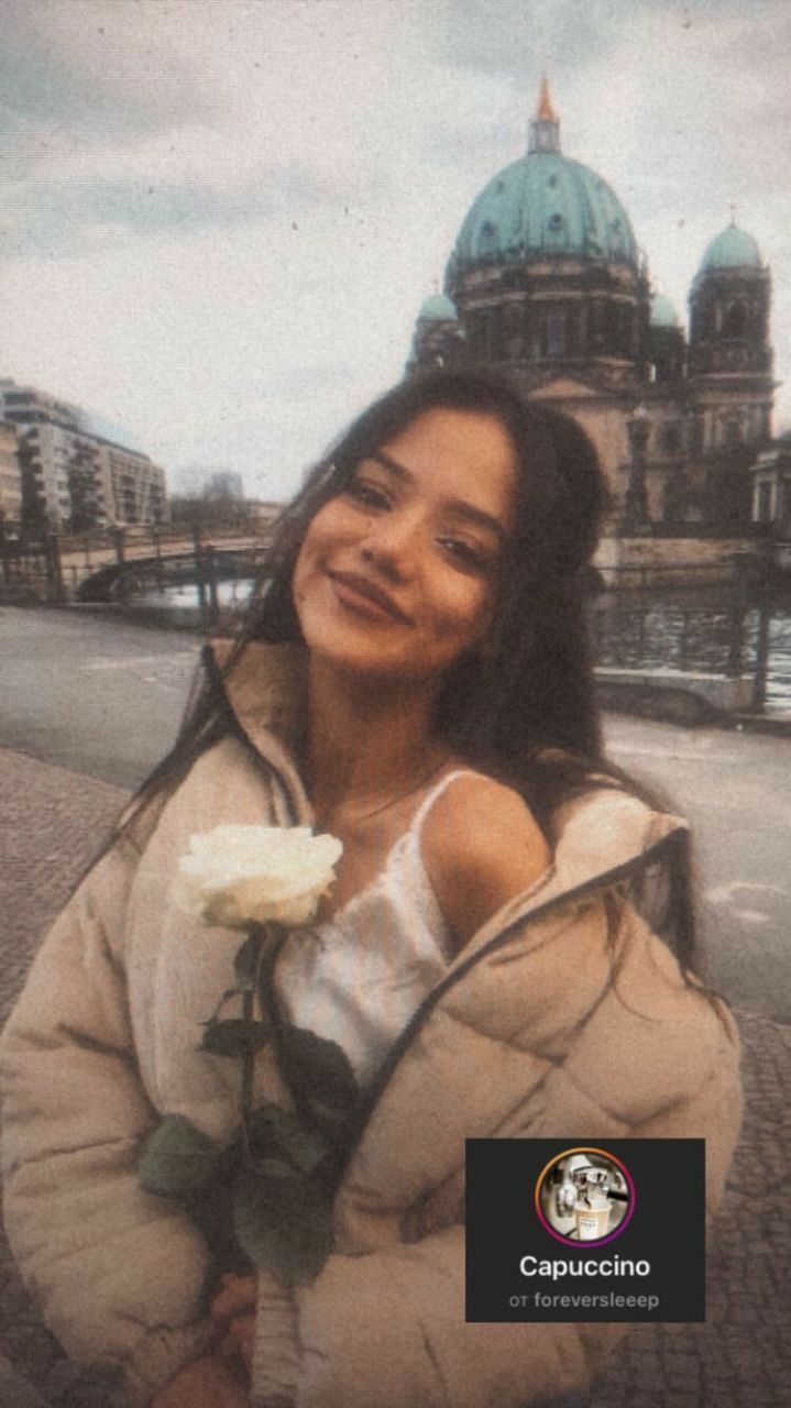 a woman holding a white rose in her right hand and looking at the camera with a building in the background