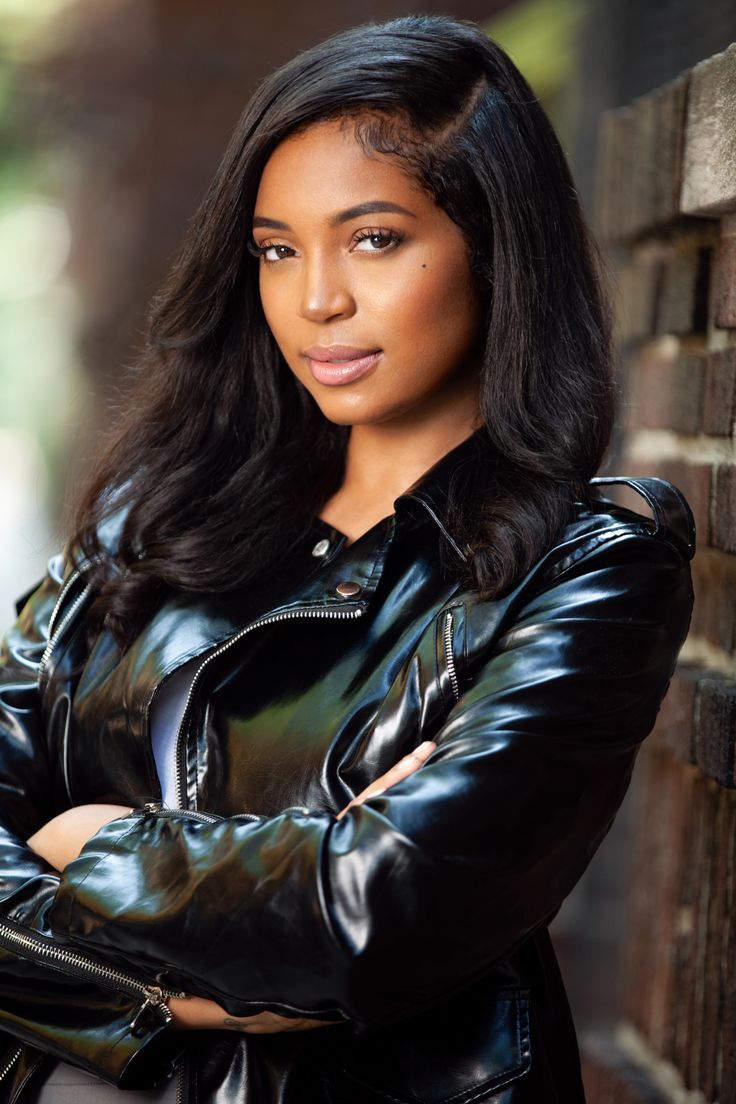 a woman wearing a black leather jacket leaning against a brick wall with her arms crossed