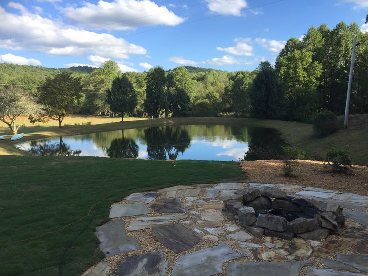 a stone fire pit in the middle of a grassy area next to a lake and trees