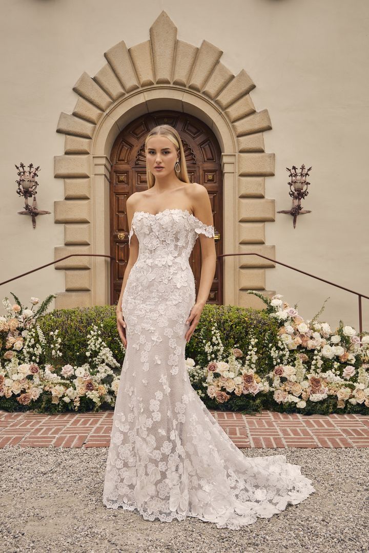 a woman standing in front of a building wearing an off the shoulder wedding dress with flowers on it