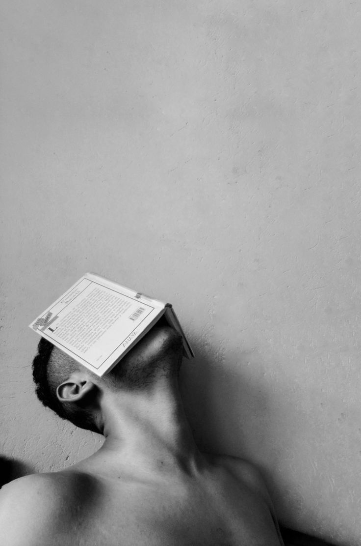 a shirtless man laying down with a book on top of his head in front of a wall