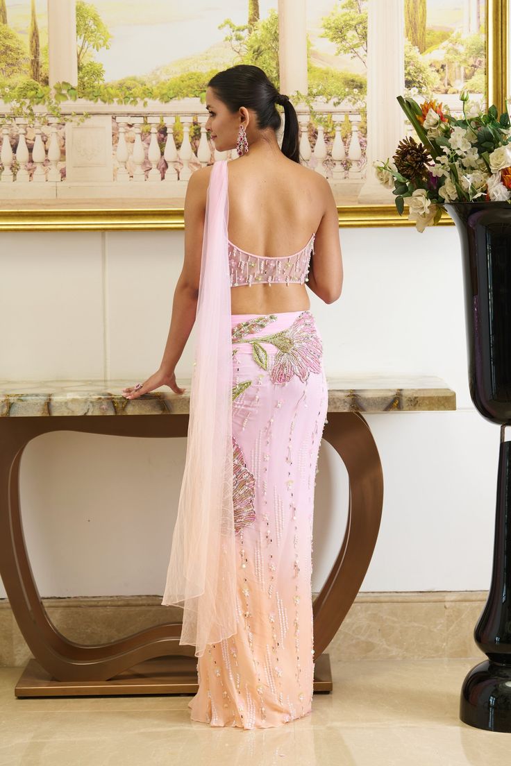 Step into the allure of pastels. The bright pink hue saree adorned with textured applique work highlighted with shiny crystals and beads crafts out a flower going all along attached with the pre-draped skirt. The shade of peach fuzz at the bottom of the silhouette brings a freshness of summer while the dainty detailing of crystal droplets all over the silhouette gives out a bling factor. Strapless bustier blouse adorned with applique work with the use of sparkling beads and crystals with an atta Pink Evening Sharara With Traditional Drape, Pink Organza Party Wear Pre-draped Saree, Evening Pink Sharara With Traditional Drape, Glamorous Pink Designer Wear Saree, Pink Party Wear Pre-draped Saree With Mirror Work, Glamorous Pink Saree For Festive Occasion, Evening Pink Saree With Resham Embroidery, Pink Tissue Silk Choli With Mirror Work, Evening Pink Traditional Drape Sharara