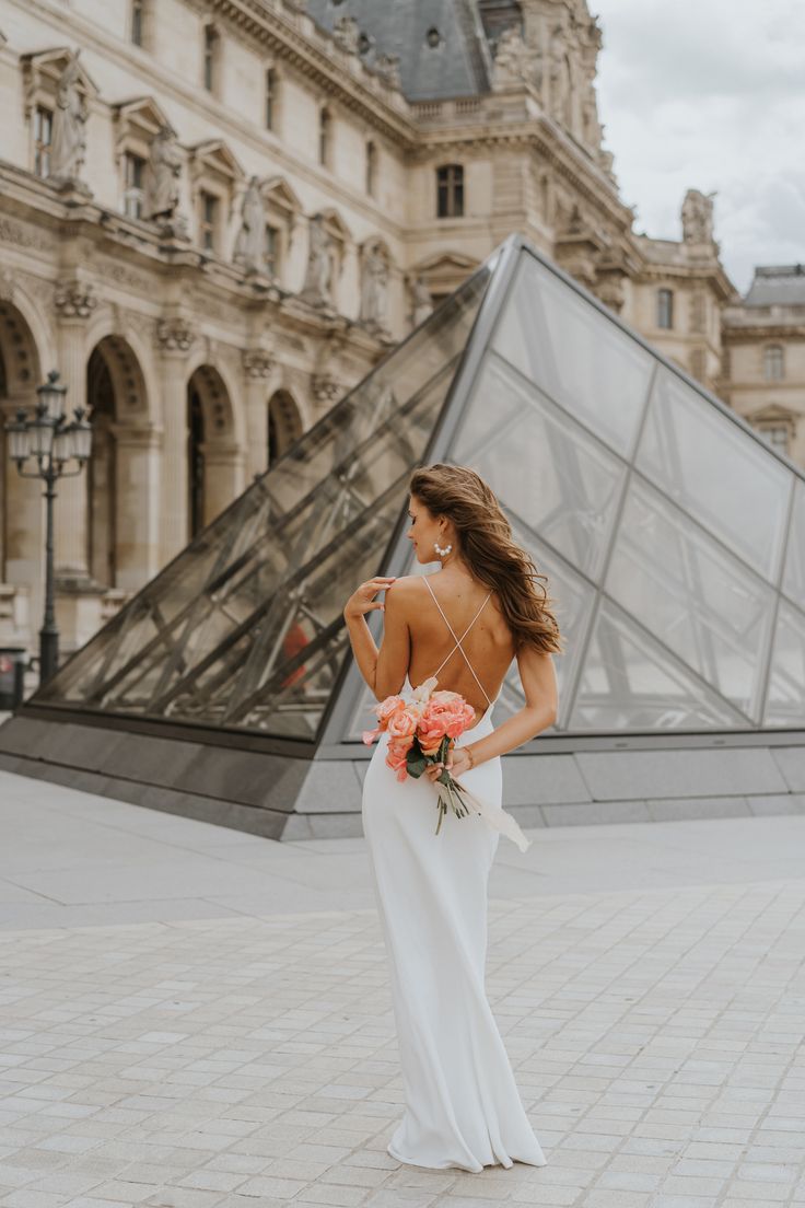 a woman standing in front of a building wearing a white dress and holding an orange bouquet