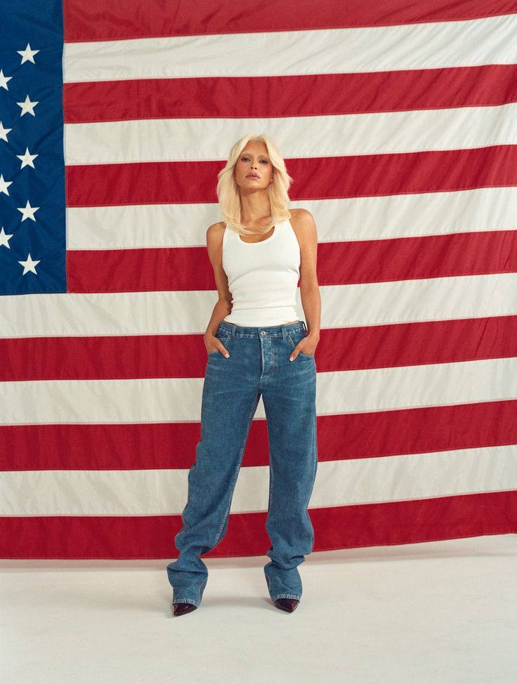 a woman standing in front of an american flag with her hands on her hips and looking at the camera