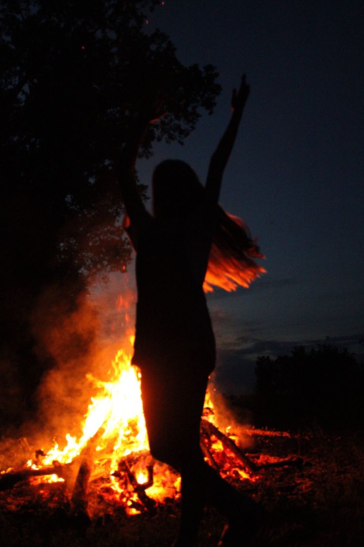 a woman standing in front of a fire with her arms up to the sky at night