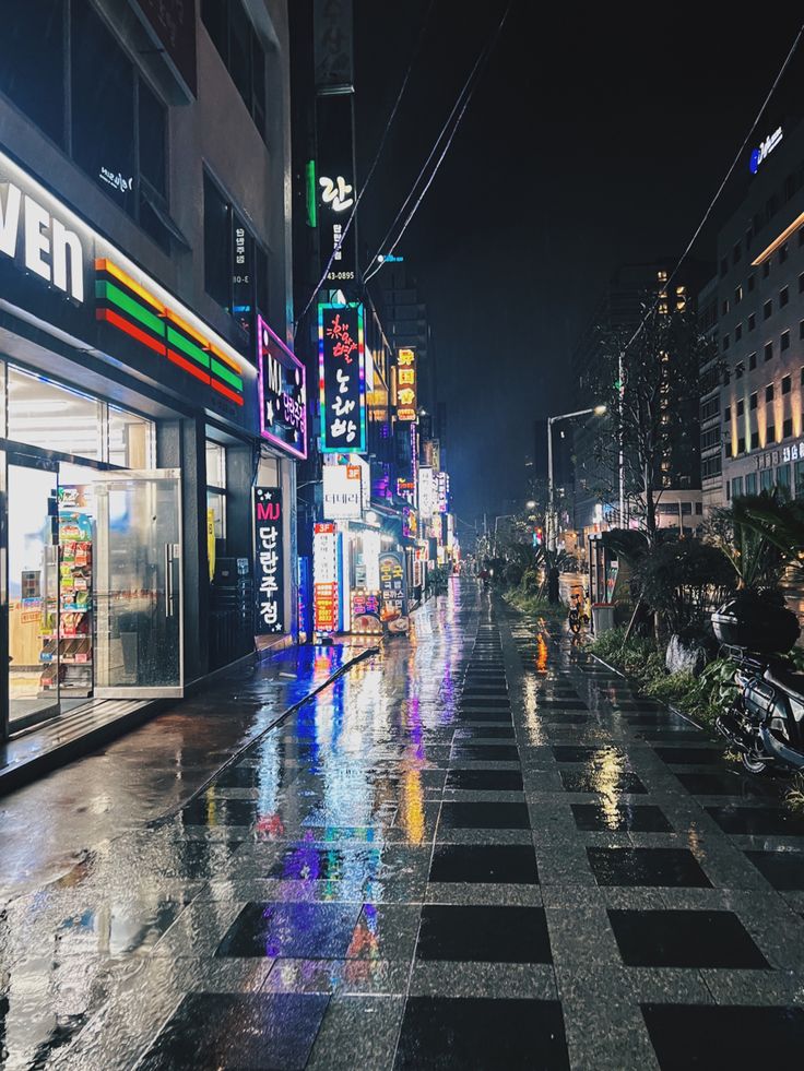 an empty city street at night with neon signs on the buildings and people walking in the rain