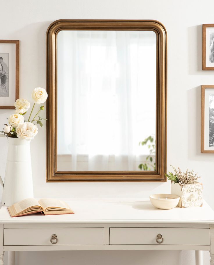 a white desk with a mirror, vase and flowers on it in front of pictures