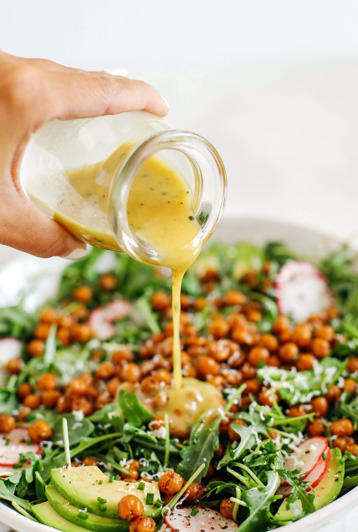 someone pouring dressing onto a salad with chickpeas and cucumbers in a white bowl