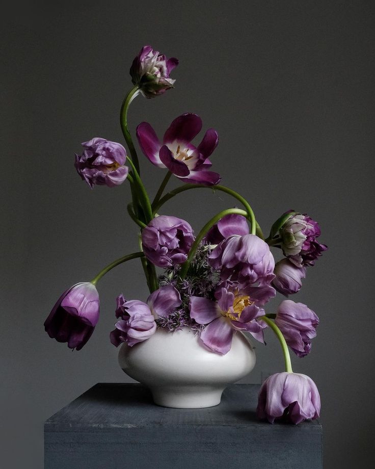 a white vase filled with purple flowers sitting on top of a wooden table next to a gray wall