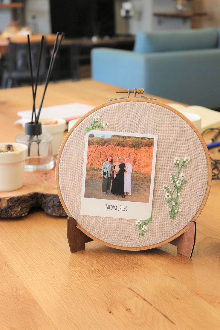 an embroidered picture frame with two women standing in front of it on a wooden table