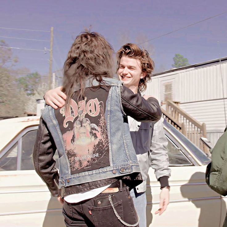 two young men are hugging each other in front of a white car and one is wearing a jean jacket