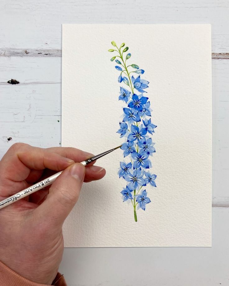 a person is painting blue flowers on white paper with watercolor pencils in their hands