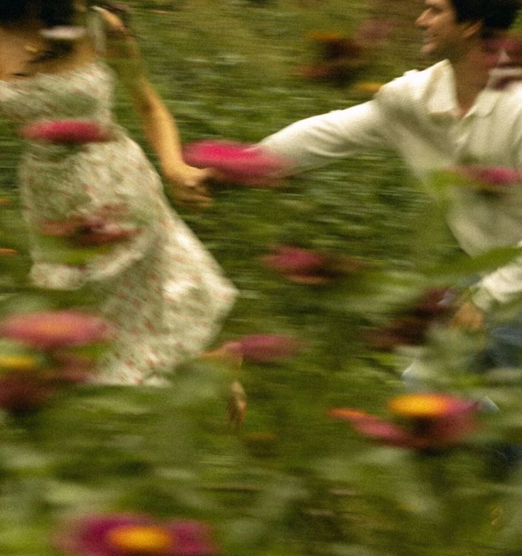 a man and woman walking through a field full of flowers with one holding the other's hand