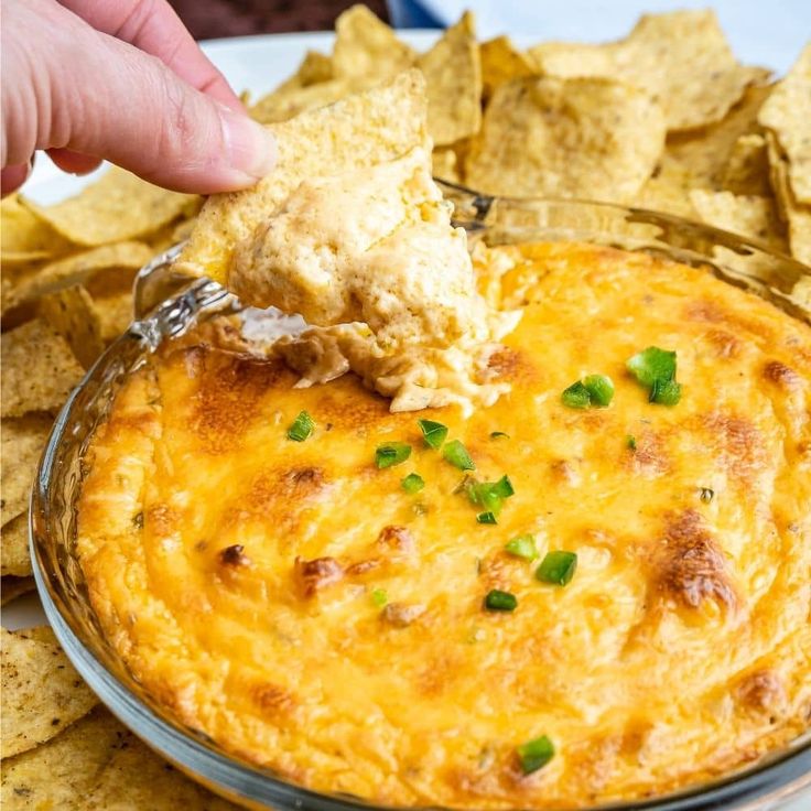 a hand dipping a tortilla chip into a bowl of cheesy dip