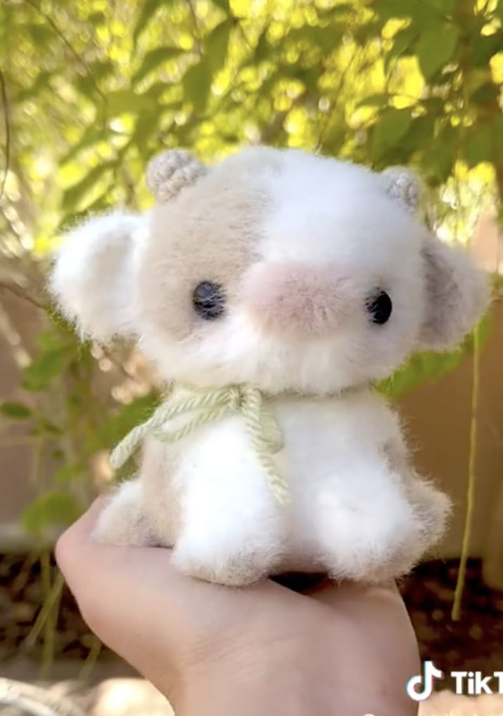 a small white teddy bear sitting on top of a persons hand in front of a tree