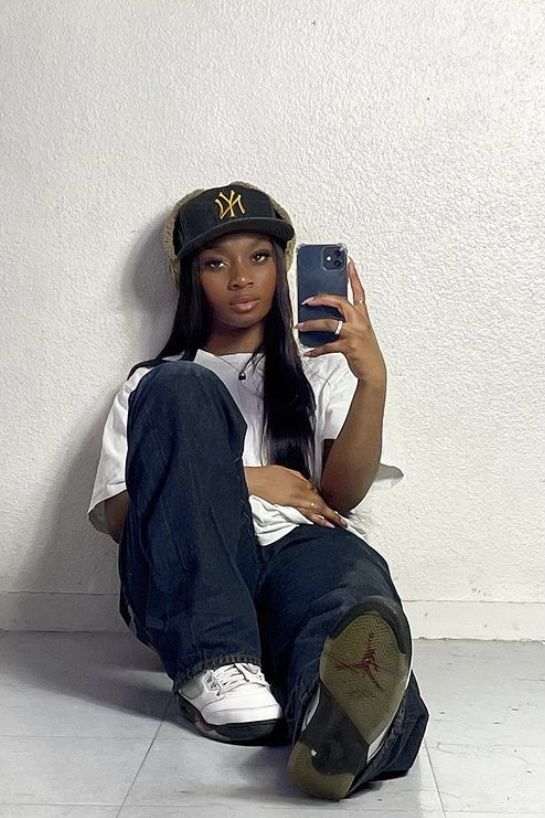 a woman sitting on the floor with her cell phone up to her ear and wearing a baseball cap