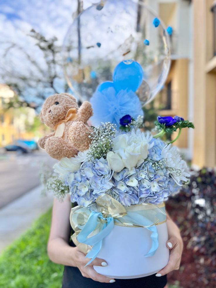 a woman holding a white vase filled with blue flowers and teddy bears on top of it