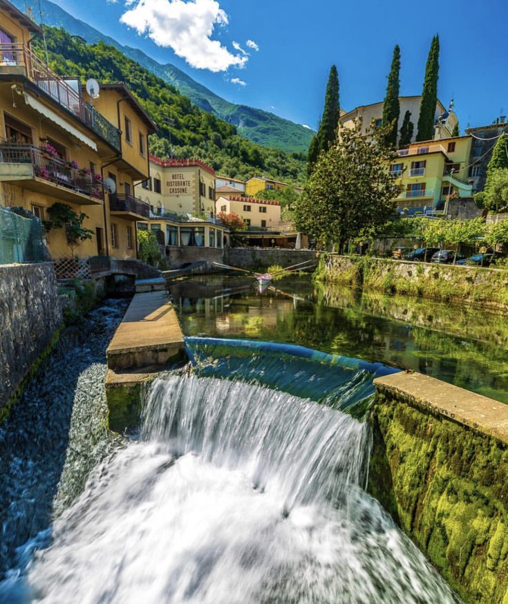 a small waterfall in the middle of a river surrounded by buildings and trees with mountains in the background