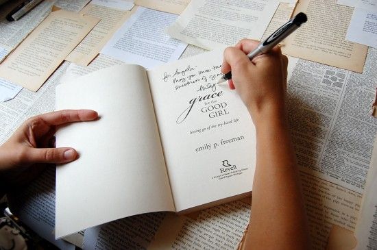 a person writing on an open book with a pen in their hand and papers scattered all over the table