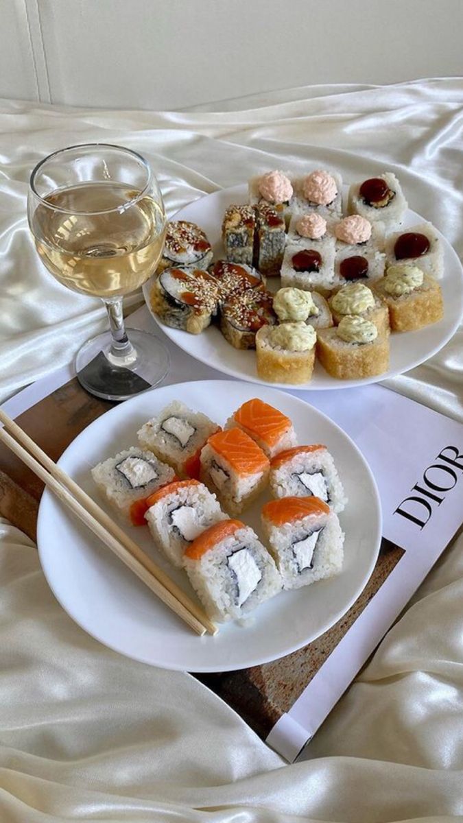 two plates with sushi and chopsticks next to a glass of white wine