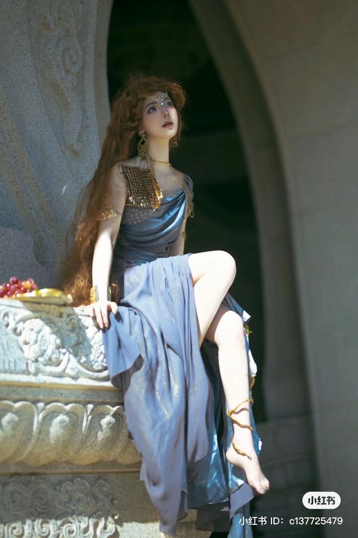 a woman with long red hair sitting on a stone wall wearing a blue dress and gold jewelry