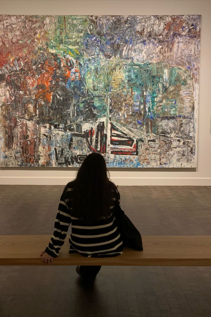 a woman sitting on top of a wooden bench in front of a large piece of art