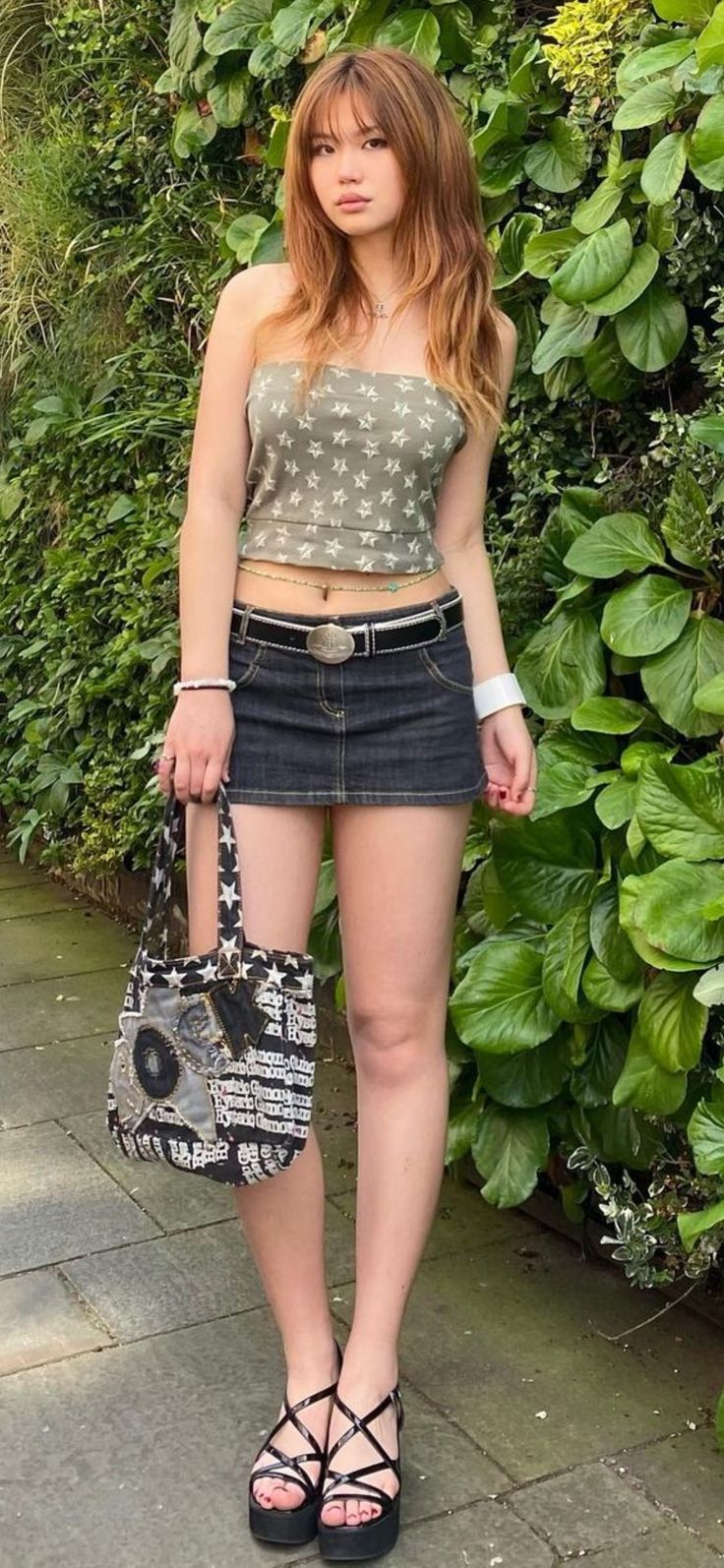 a woman standing in front of a bush holding a purse and looking at the camera