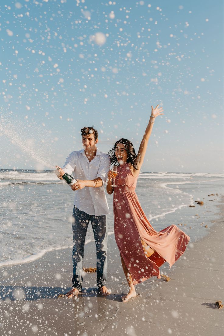 a man and woman standing on top of a beach next to the ocean holding wine bottles