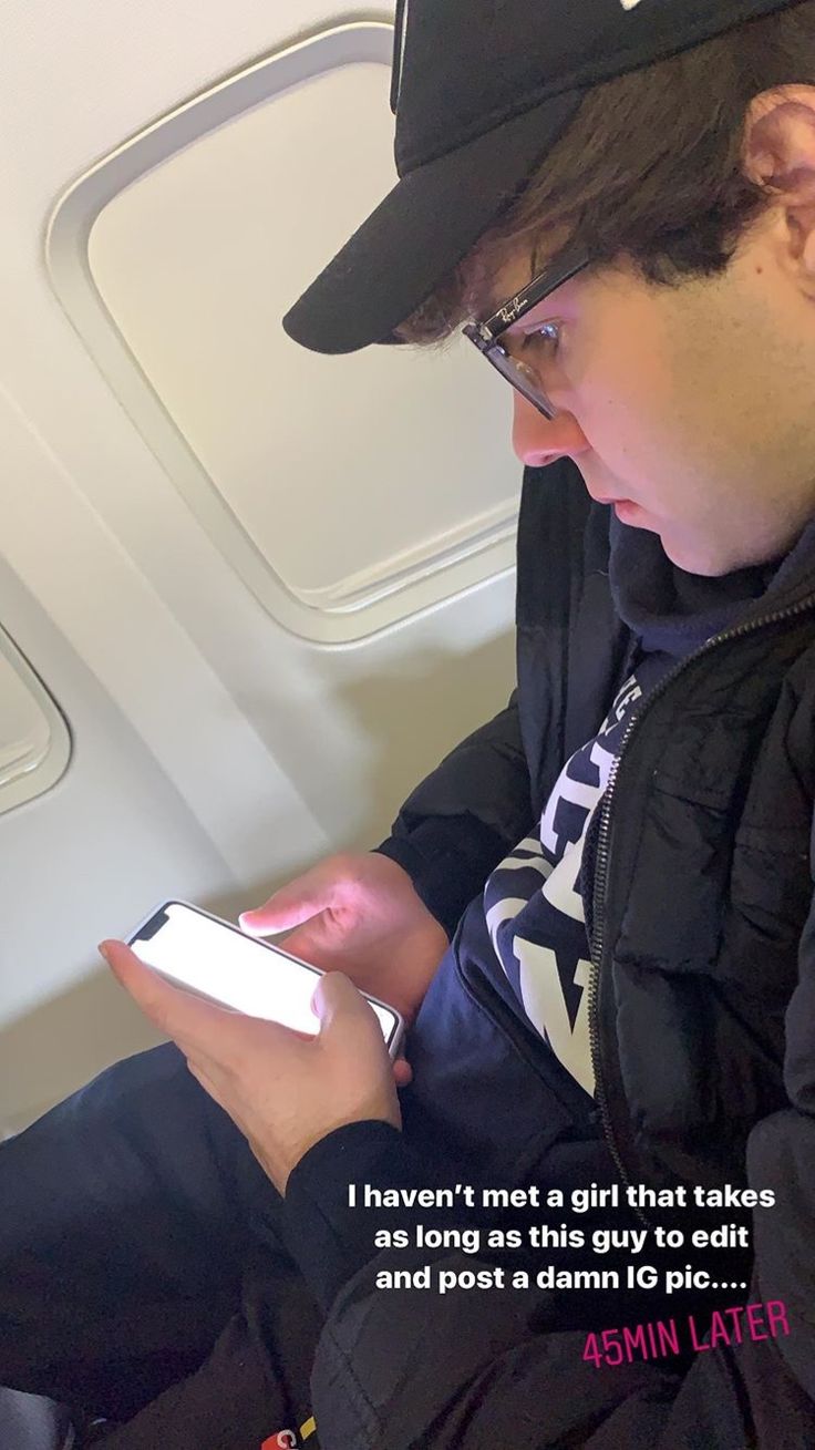 a man sitting on an airplane looking at his cell phone while wearing a baseball cap