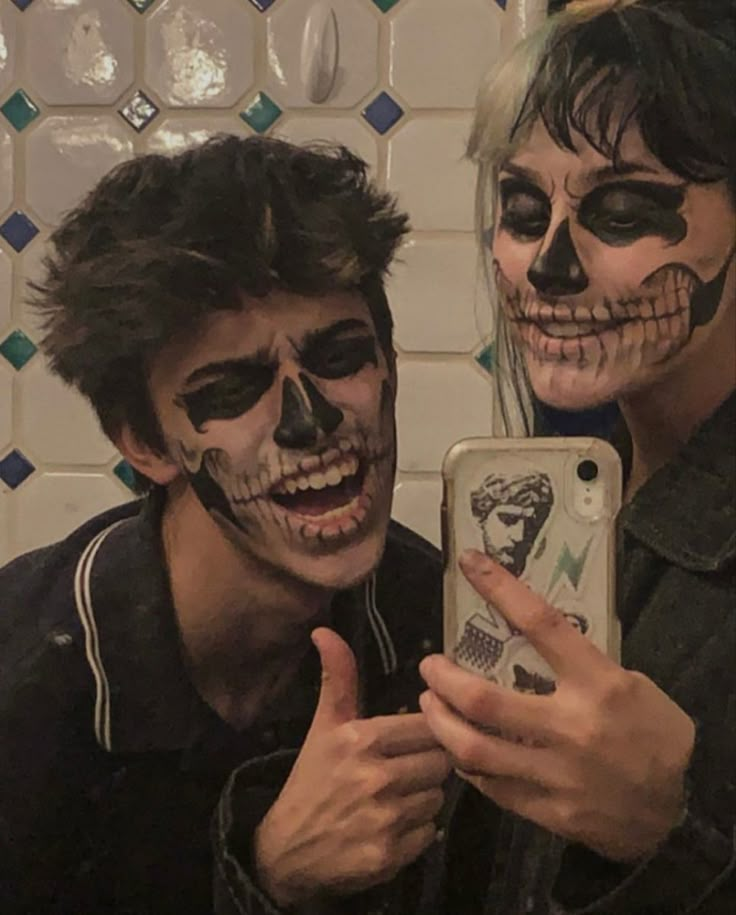 two people with face paint taking a selfie in front of a tiled wall and holding a cell phone