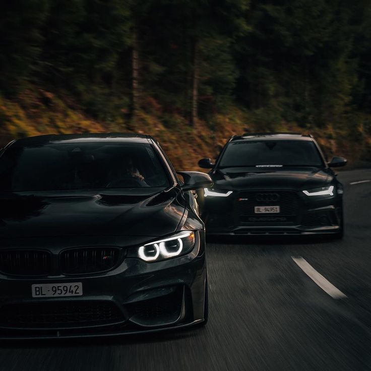 two black cars driving down the road next to each other in front of some trees