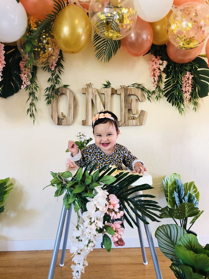 a baby sitting in a chair with flowers and balloons behind her, smiling at the camera