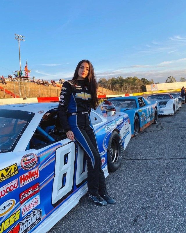 a woman leaning on the back of a race car in a parking lot with other cars