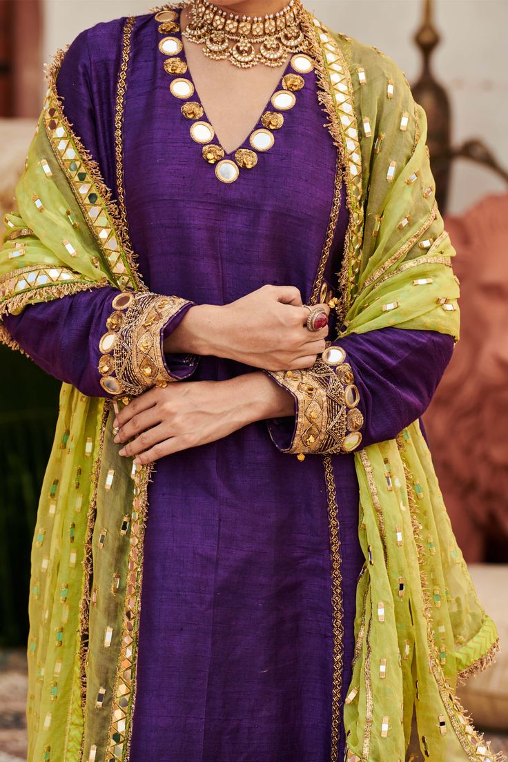 A striking ensemble, this purple raw silk sharara set showcases exquisite mirror work on the neckline and sleeves of the tunic, exuding elegance and charm. Paired with matching pants and a contrasting lime green organza dupatta, this outfit beautifully balances richness and vibrancy, making it a perfect choice for festive celebrations and special events. Unstitched Purple Raw Silk Kurta, Purple Palazzo Set With Dabka Work And Straight Kurta, Purple Raw Silk Salwar Kameez For Festivals, Festival Purple Raw Silk Salwar Kameez, Designer Raw Silk Purple Salwar Kameez, Designer Purple Raw Silk Salwar Kameez, Purple Chanderi Kurta With Mirror Work, Designer Wear Purple Raw Silk Salwar Kameez, Semi-stitched Purple Palazzo Set With Mirror Work